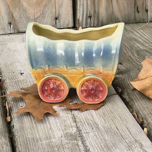 May include: A ceramic wagon planter with a rectangular opening, painted in shades of blue, yellow, and orange. The planter has two red wheels and sits on a wooden surface with a dried leaf.