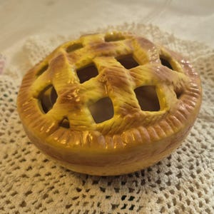 May include: A ceramic pie-shaped container with a lattice top. The container is painted in shades of yellow and brown, with a glossy finish. The pie has a round shape and is sitting on a white, textured surface.