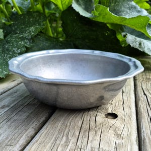 May include: A round, silver-toned metal bowl with a scalloped, wavy rim. The bowl has a matte, textured finish and a shallow, wide basin. The design is simple and functional, featuring a consistent metallic gray color throughout the entire piece.