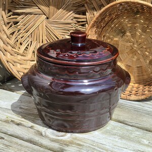 May include: A dark brown glazed ceramic crock with a matching lid, featuring a rounded body, two side handles, and embossed floral and wavy line patterns around the exterior. The pot has a glossy finish and a small knob on the center of the lid.