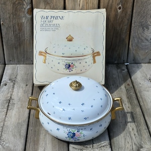 May include: A 5-quart Dauphine Dutch oven made of porcelain on steel with brass trim. The white pot and lid feature a small blue floral pattern and a larger floral cluster on the front. It includes a matching box with product text.