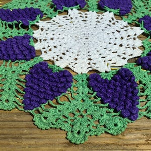 Puede incluir: Un tapete de ganchillo hecho a mano con un diseño central de estrella blanca, rodeado de hojas verdes y racimos de uvas moradas. Los intrincados detalles y los colores vibrantes crean una pieza decorativa.