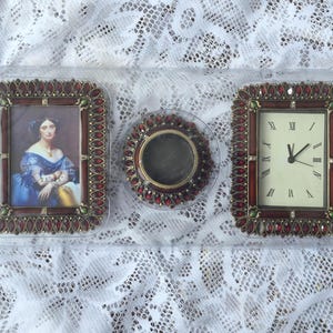 May include: A set of three decorative items in a clear plastic tray, including a rectangular picture frame with a portrait, a circular candle holder, and a rectangular clock with Roman numerals. Each piece features a dark red border with red gems.