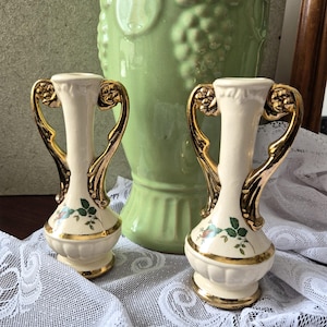 May include: Two cream-colored vintage bud vases with gold handles and trim. Each vase features a floral design with green leaves and red accents. A large green vase is in the background, along with a white lace tablecloth.