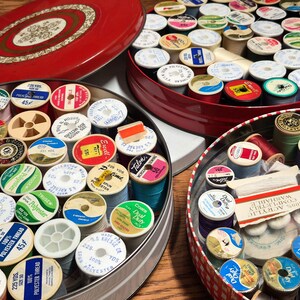 May include: A collection of vintage thread spools in various colors and brands, neatly arranged inside several red and silver tins. The spools are labeled with text indicating thread type, yardage, and brand names. The tins are open, showcasing the assortment of sewing supplies.