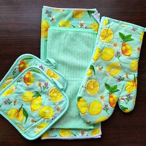 May include: A kitchen set featuring a light green background with a lemon pattern. The set includes an oven mitt, two pot holders, and two towels. The lemons are yellow with green leaves and white flowers. The set is designed for kitchen use.