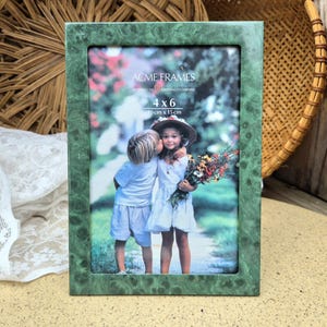 May include: Green picture frame with a marble effect, holding a 4x6 inch photo of two children. The frame has the text "ACME FRAMES" and "4 x 6" printed on it. The photo shows two children, one kissing the other on the cheek.