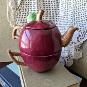 May include: A whimsical, apple-shaped teapot in shades of burgundy, with a green leaf and a brown stem. The teapot has a matching cup base, and brown handles and spout. The teapot is sitting on top of a stack of books.