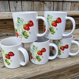 May include: Six white ceramic mugs stacked on a wooden surface. Each mug features a design of red strawberries, green leaves, and small blue and white flowers. The mugs have large, curved handles.