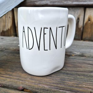 May include: White ceramic mug with the word "ADVENTURE" in black lettering. The mug has a curved handle and a slightly irregular shape. The mug is sitting on a wooden surface.