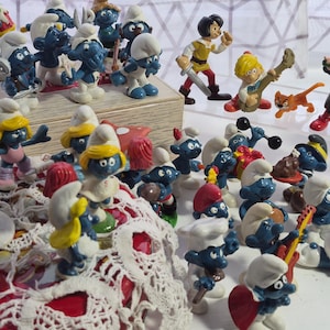 May include: A collection of Smurf figurines in various poses, with blue skin and white hats. Some figurines are playing musical instruments, while others are engaged in different activities. Also included are figurines of other cartoon characters.