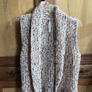 May include: A sleeveless, open-front vest in a white knit fabric with colorful flecks. The vest hangs on a clear plastic hanger against a wooden background. The collar is a shawl style.
