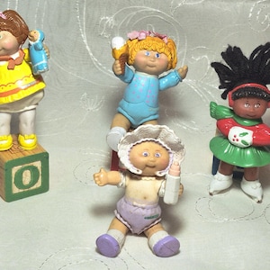 May include: Four Cabbage Patch Kids figurines. One doll stands on a wooden block with the number 0. The dolls are wearing various outfits in yellow, blue, red, and green. One doll is holding a bottle.