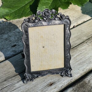 May include: A rectangular picture frame with a dark metallic finish and a textured surface. The frame features ornate rose motifs at the top and bottom corners, with a beaded inner border surrounding a plain, light-colored rectangular insert.