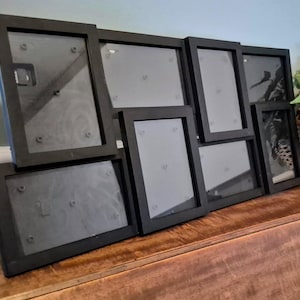 May include: A multi-photo black picture frame with various sized openings. The frame is made of wood and is displayed on a wooden surface. A woven basket with greenery is in the background.
