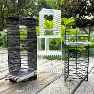 May include: Two black metal wire racks with horizontal slots for media storage and one clear acrylic storage unit with stacked compartments, all designed for organizing compact discs or similar flat items in a vertical arrangement.