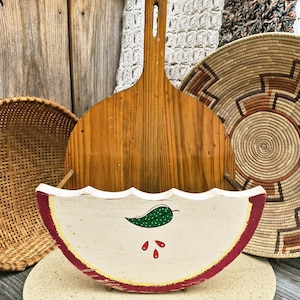 May include: A wooden pizza peel and a decorative, hand-painted, watermelon-shaped wall pocket. The wall pocket is white with a red border and features a green pepper and red seed details. Woven baskets and a round stone base are in the background.
