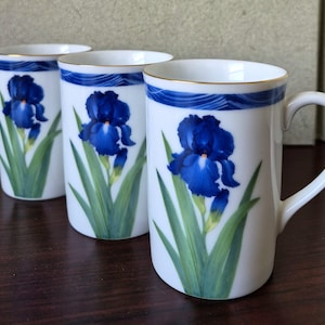 May include: Three white ceramic mugs with a blue iris flower design. Each mug features a gold rim and a blue wave pattern near the top. The iris is painted in shades of blue and green.