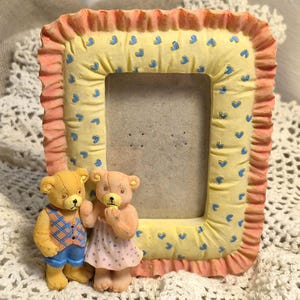 May include: A rectangular photo frame with a yellow background adorned with blue hearts and a ruffled peach border. Two teddy bear figurines stand in front of the frame. One bear wears a vest and shorts, the other a dress.
