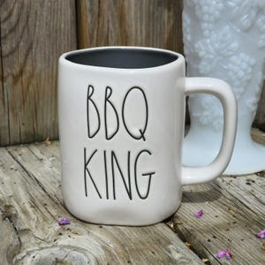 May include: White ceramic mug with the words "BBQ KING" in black lettering. The mug has a dark gray interior and a curved handle. The mug is on a wooden surface.