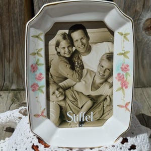 May include: White ceramic picture frame with a gold trim and floral accents. The frame holds a 4"x6" photo and features the brand name "Stiffel". The frame has a rectangular shape with a scalloped edge and is decorated with pink roses and green leaves.