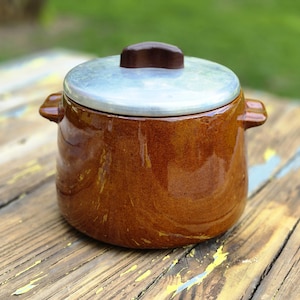 May include: A brown ceramic pot with a silver lid and a dark brown handle. The pot has two small handles on either side. The pot is sitting on a weathered wooden surface.