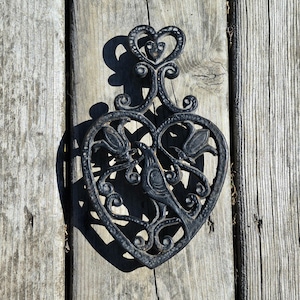 May include: Black cast iron trivet in a heart shape, featuring a bird and floral design. The trivet has a decorative handle with a heart-shaped detail. The trivet is placed on a weathered wooden surface.