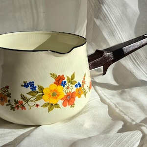 May include: Cream-colored enamelware saucepan with a floral design featuring yellow, orange, and blue flowers. The pot has a dark brown handle and a black rim. A vintage kitchenware item.