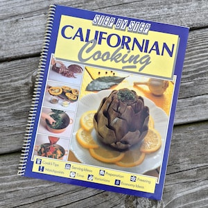 May include: A spiral-bound cookbook titled "Step by Step Californian Cooking" with a blue cover. The cover features images of food, including an artichoke with orange slices. The book is on a weathered wooden surface.