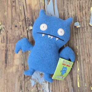 May include: A blue plush toy shaped like a bat with large white eyes, small fangs, and wings. The toy has a tag that reads "Little Uglys Ice-Bat" and "Uglydoll". The toy is on a weathered wooden surface.
