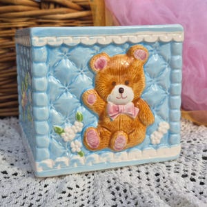 May include: A light blue ceramic box with a quilted pattern and a raised teddy bear design. The bear is brown with a pink bow tie. White flowers and trim add detail to this decorative box.
