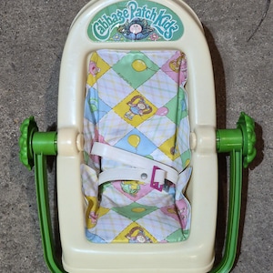 May include: A cream-colored plastic Cabbage Patch Kids doll carrier featuring a green handle and a fabric insert with a diamond pattern, yellow balloons, and doll illustrations. It includes a white safety strap with a pink plastic buckle.