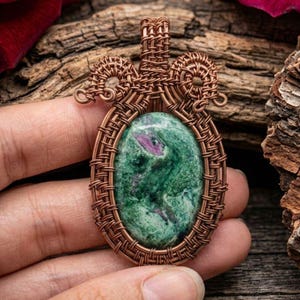 Puede incluir: Un colgante ovalado con una piedra preciosa de zoisita verde y magenta engastada en un intrincado alambre de cobre. El trabajo en metal forma un marco decorativo alrededor de la piedra con una anilla trenzada en la parte superior.