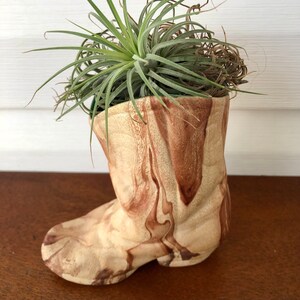 May include: A ceramic boot planter with a marbled beige and brown pattern, containing a green air plant and dried moss. The boot planter is a unique home decor item.
