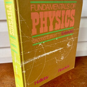 May include: A vintage textbook titled "Fundamentals of Physics" in a mustard yellow cover. The title is in large orange letters with green accents. The book is a second edition, extended, by Halliday and Resnick.