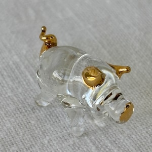 May include: A clear glass pig figurine with gold accents. The pig has a curled tail, a snout with gold trim, and a gold eye.