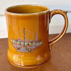 Puede incluir: Una taza de cerámica marrón con borde y asa dorados. La taza presenta una colorida ilustración de la Columna de Nelson en Trafalgar Square. El texto "NELSON'S COLUMN TRAFALGAR SQUARE" está impreso debajo. Una taza de recuerdo vintage.