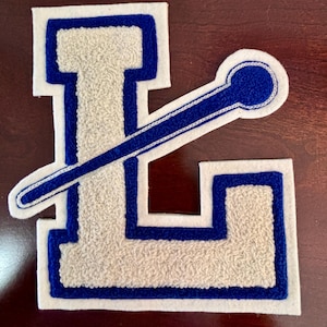 May include: A varsity-style letter L patch featuring a textured cream-colored center with a bold blue border and a white felt backing. A diagonal blue line with a circular end extends across the letter, creating a layered, embroidered appearance.