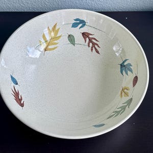 May include: A round, cream-colored ceramic bowl with a speckled finish. The interior features a decorative pattern of scattered autumn leaves in shades of yellow, brown, green, and blue, arranged in a circular formation around the center.