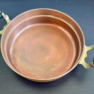 Op de afbeelding: Een ronde koperen pan met een gepolijst binnenoppervlak. De pan is voorzien van twee koperen zijhandgrepen, elk uitgerust met een zwarte cilindrische greep. De metalen constructie vertoont een warme metaalkleur over het gehele bekken.