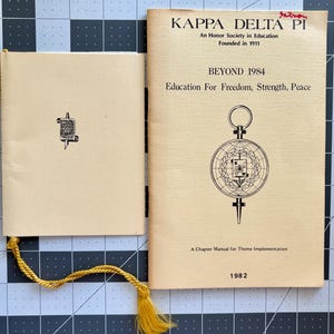 May include: A yellow and white pamphlet with the title "Kappa Delta Pi" and the text "An Honor Society in Education Founded in 1911" and "Beyond 1984 Education For Freedom, Strength, Peace". The pamphlet also includes a black and white illustration of a compass with a globe and a scroll.