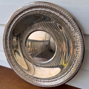 May include: A silver-colored, round serving dish with a decorative border. The dish has a reflective surface, with a central indentation. The rim features an ornate, repeating pattern. The dish is set on a wooden surface.