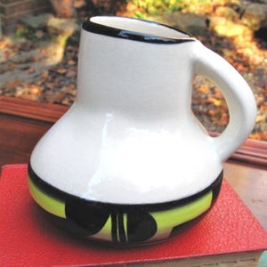 May include: A white ceramic jug with a black rim and handle. The base has a design with black and yellow accents. The jug is on a red surface, with a blurred background of foliage.