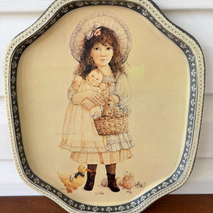 Könnte beinhalten: Ein vintage Serviertablett aus Metall mit erhöhtem Rand und einem gewellten Design. Das Tablett zeigt eine farbenfrohe Illustration eines jungen Mädchens, das eine Puppe und einen Korb mit Hasen hält. Das Mädchen trägt ein weißes Kleid mit einer Spitzenborte und eine weiße Haube. Das Tablett hat einen schwarz-weißen floralen Rand.