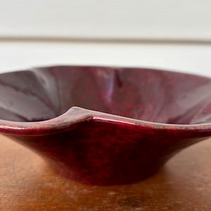 Puede incluir: Un cuenco de cerámica burdeos con un borde ondulado único. El cuenco tiene un acabado brillante y una forma acampanada, mostrando un rico tono rojo oscuro. El cuenco es probablemente hecho a mano y adecuado para uso decorativo o funcional.