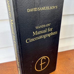 May include: A black leather-bound book with gold lettering titled "'HANDS-ON' Manual for Cinematographers" by David Samuelson. The book has a gold "F" in a circle on the front cover.