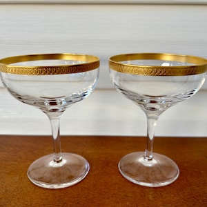 Puede incluir: Dos copas de champán de vidrio transparente con bordes dorados. Los bordes tienen un patrón decorativo.