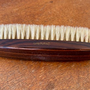 May include: A wooden brush with light-colored bristles. The dark brown handle has a curved shape and the text "Trophy-Tastic" is printed on the handle. The brush is designed for grooming.