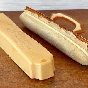 Op de afbeelding: Twee vintage schoenpoetssets. Eén is een rechthoekige, lichtgele doos met handgeschreven tekst. De andere is een lichtbeige schoenborstel met een bruin handvat. Beide liggen op een houten oppervlak.