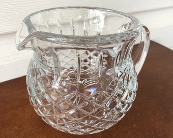 May include: A clear glass pitcher with a diamond pattern. The pitcher has a handle and a spout.